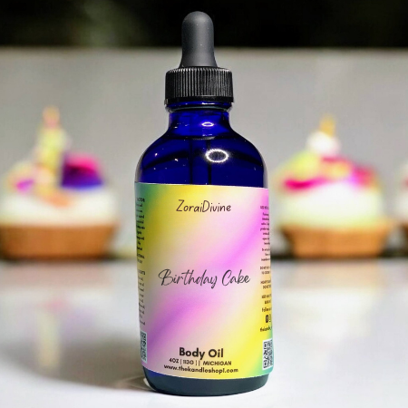 Bottle of ZoraiDivine Birthday Cake body oil with colorful label, blurred cupcakes in the background