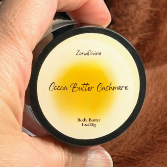 Hand holding a container of ZoraiDivine  Cocoa Butter Cashmere body butter against a brown fabric background.