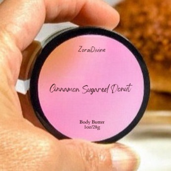 Hand holding a container of ZoraiDivine  Cinnamon Sugared Donut body butter with a blurred background.