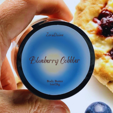 Hand holding a container labeled 'Blueberry Cobbler' body butter next to a blueberry cobbler dessert.