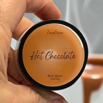 Hand holding a container of body butter labeled 'Hot Chocolate' with a blurred background