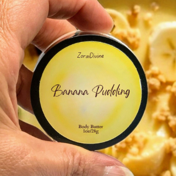 Hand holding a container labeled 'Banana Pudding' body butter with a dessert in the background
