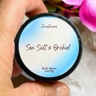 Hand holding a container of ZoraiDivine  Sea Salt & Orchid body butter with flowers in the background
