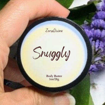 Hand holding a container of 'Snuggly' body butter with green leaves and purple flowers in the background