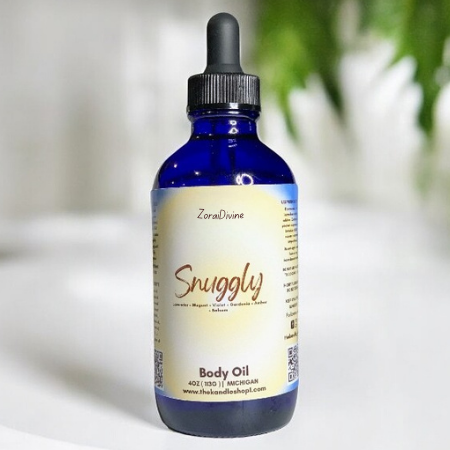 Bottle of Snuggly body oil on a white surface with a blurred green plant in the background