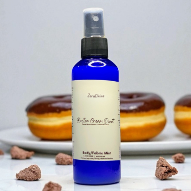 Body mist Boston cream donut on a white countertop with donuts & chocolate pieces around the bottle.